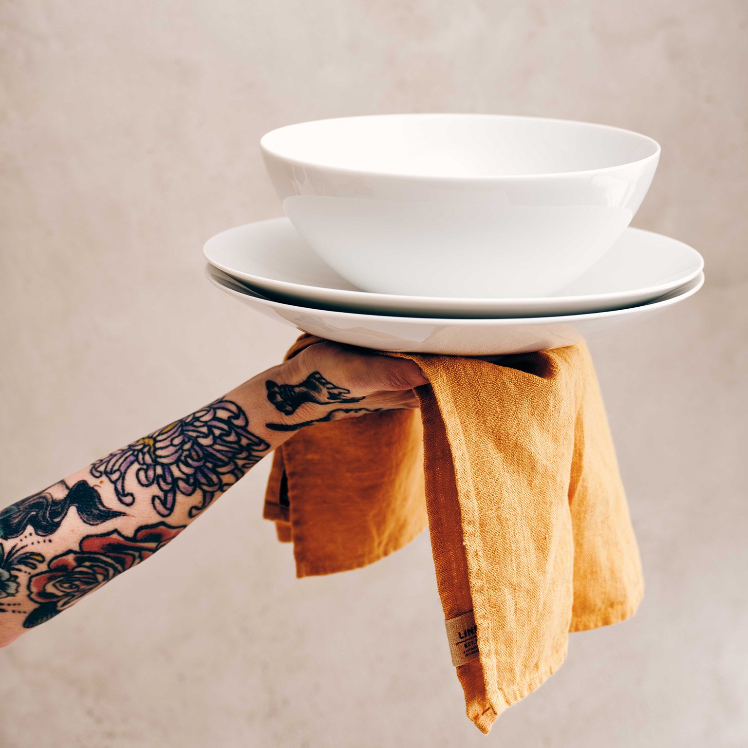 A tattooed arm on a napkin holds the TAC Allround bowl stacked on TAC Allround plate in the air.